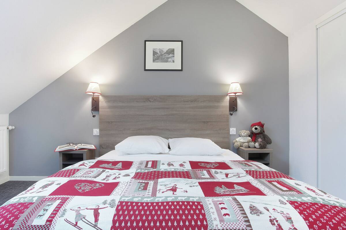 Ein Schlafzimmer mit schrägen Wänden, einer gesteppten Bettdecke, zwei Nachttischlampen, einem Teddybären und einem gerahmten Bild an der Wand.