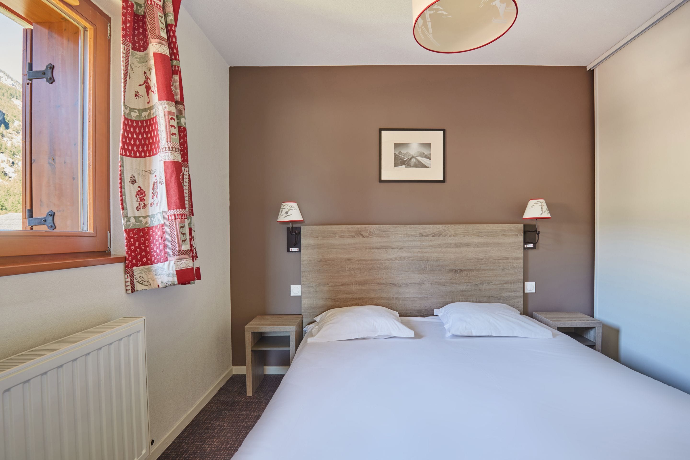 A bedroom with a wooden headboard, a bed with white linens, two bedside lamps, a framed picture on the wall, and a window with a curtain.