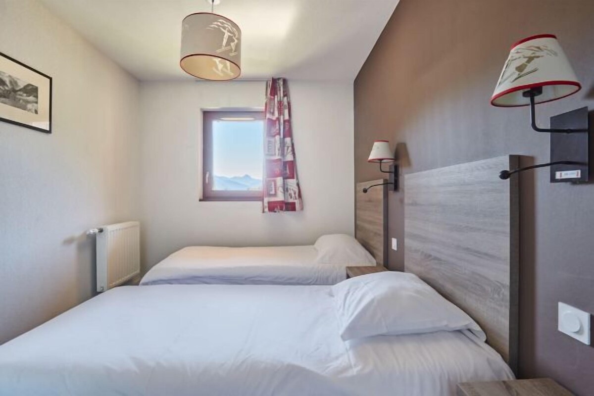 A hotel room with two beds, a window with a view, a radiator, and a lamp on the wall.