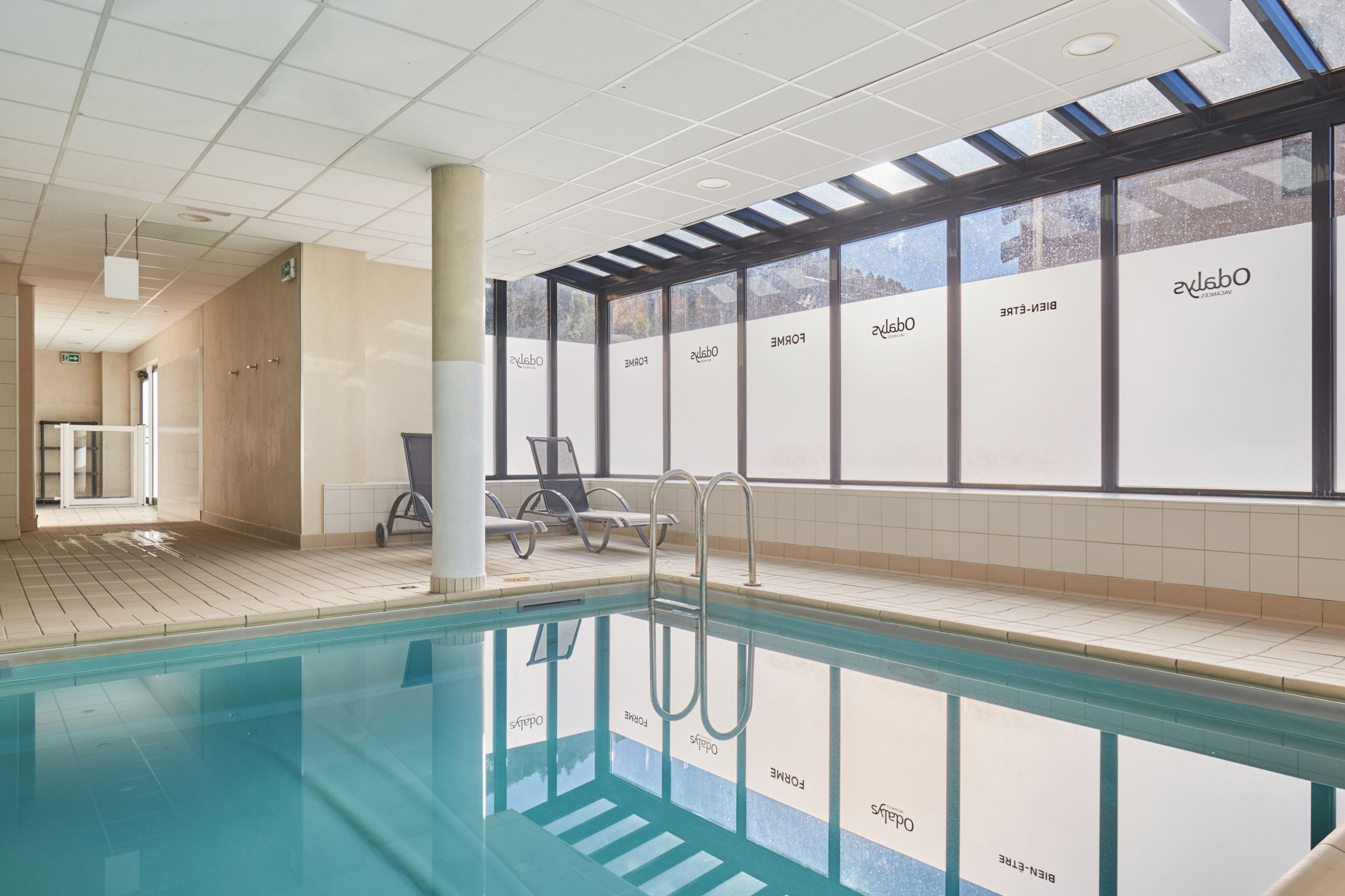 A swimming pool with clear glass walls, a tiled floor, and a ceiling with recessed lighting.