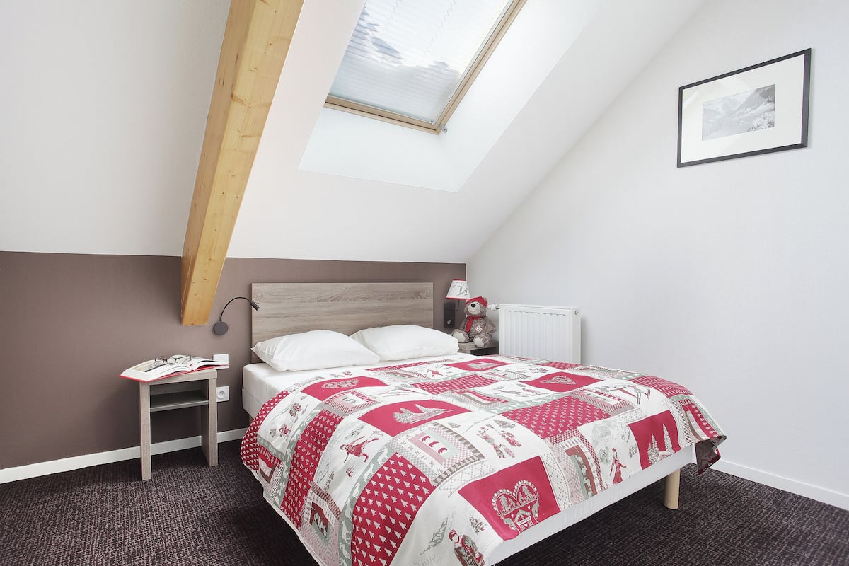 Ein Schlafzimmer mit schrägen Wänden, einem Bett mit einer rot-weißen Steudecke, einem Nachttisch, einer Lampe und einem gerahmten Bild an der Wand.