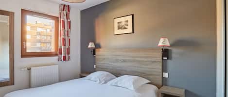 A bedroom with a bed, a window with curtains, a radiator, a wall-mounted picture, and two bedside lamps.