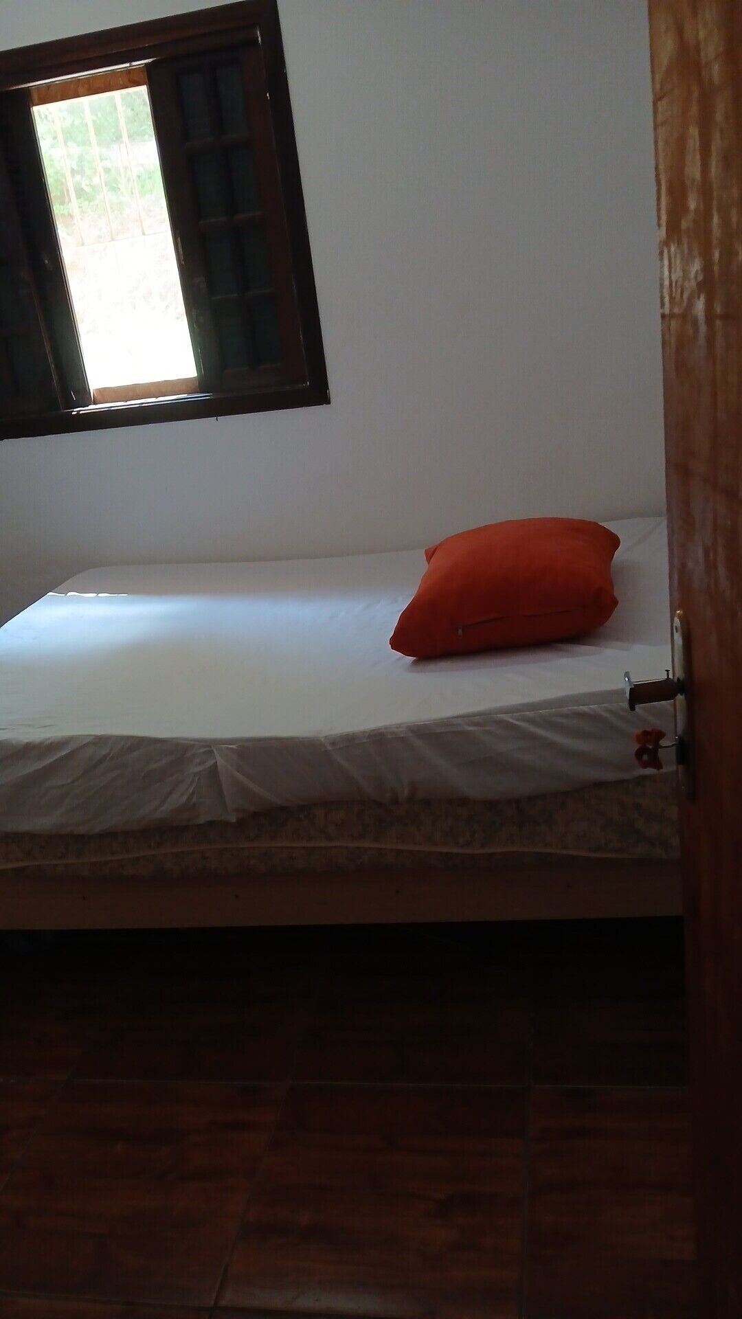 Un lit simple avec un matelas blanc et un oreiller rouge, une fenêtre avec des stores et un plancher en bois.