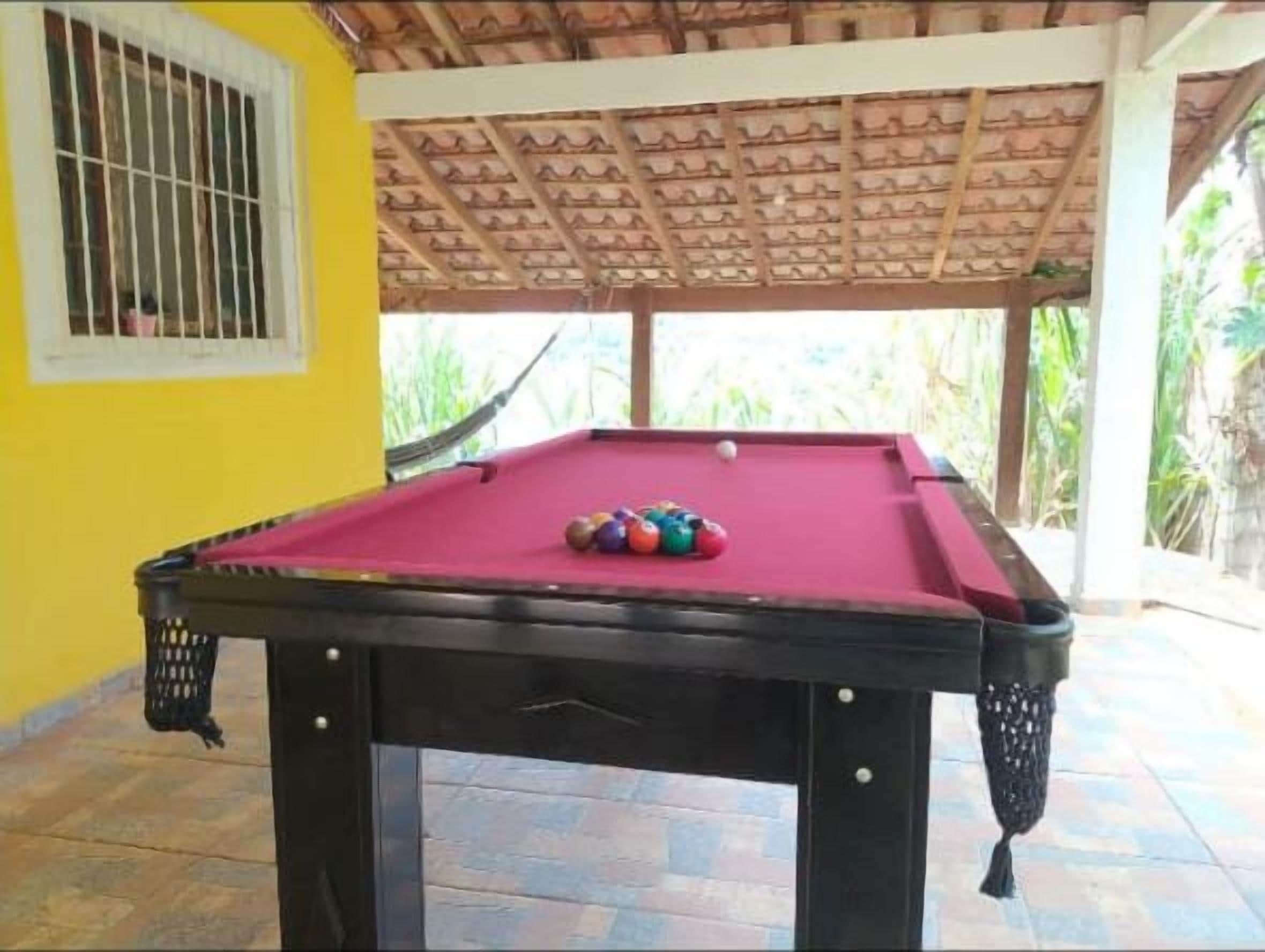 Uma mesa de bilhar com superfície vermelha e bolas de bilhar, com uma parede amarela ao fundo e uma estrutura de telhado de madeira.