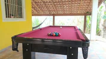 Uma mesa de bilhar com superfície vermelha e bolas de bilhar, com uma parede amarela ao fundo e uma estrutura de telhado de madeira.