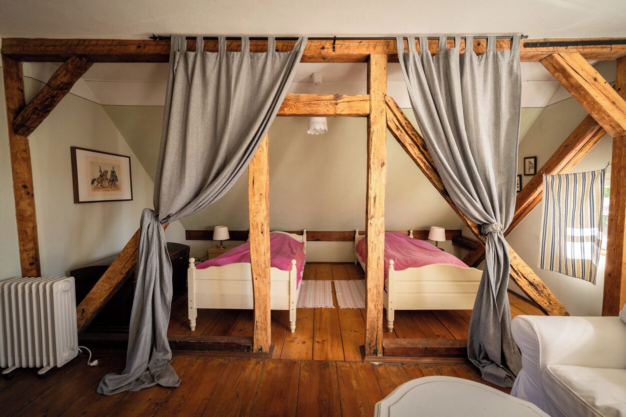 Una habitación con vigas de madera, dos camas con ropa de cama rosa, cortinas grises, un radiador y una silla blanca.
