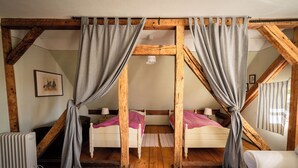 A room with wooden beams, two beds with pink bedding, grey curtains, a radiator, and a white chair.