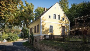 A two-story yellow building with a stone foundation, a wooden fence, and a paved road in front.