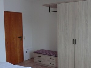 A bedroom with a wooden door, a wardrobe with two doors, a bed, and a small bench with drawers.