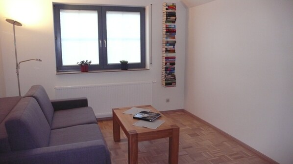 A living room with a sofa, a coffee table with books, a window with blinds, and a bookshelf.