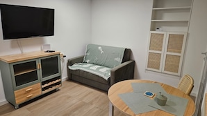 A living room with a sofa, a TV, a wooden cabinet, a dining table, and a window with blinds.
