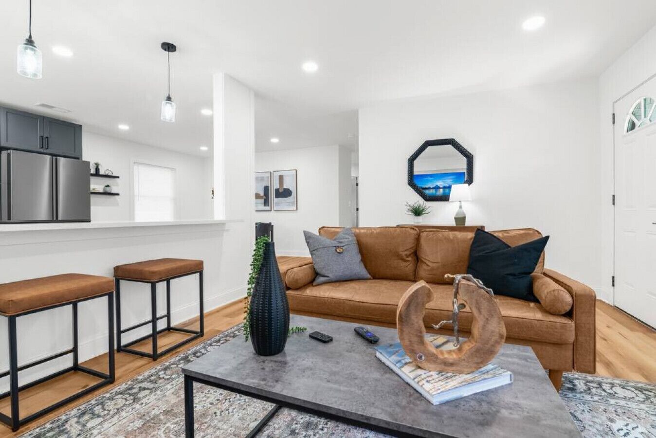 A modern living room with a brown leather sofa, two wooden bar stools, a coffee table with decorative items, and a kitchen area with stainless steel appliances.