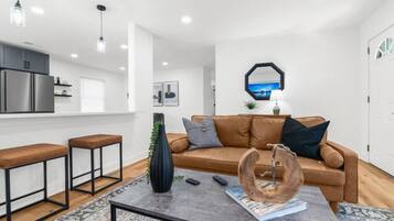 A modern living room with a brown leather sofa, two wooden bar stools, a coffee table with decorative items, and a kitchen area with stainless steel appliances.