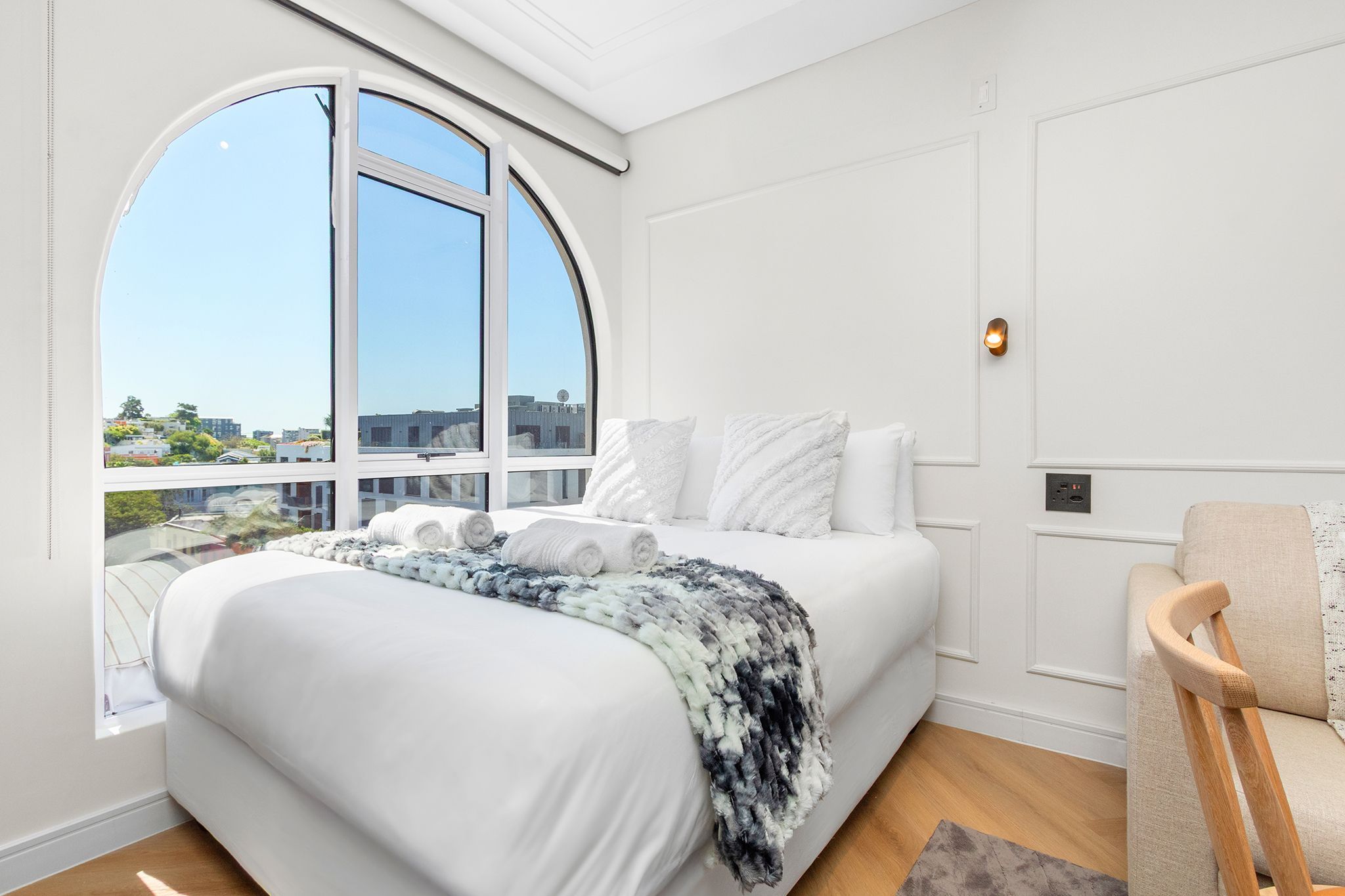 A modern bedroom with a large window, a bed with white linens, a grey and white throw blanket, and a wooden chair.