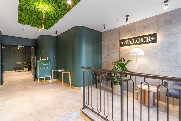 A reception area with a sign reading 'The Valour,' a green wall, a staircase with a metal railing, and a potted plant.