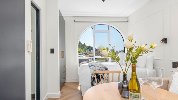 A modern hotel room with a large arched window, a bed with white linens, a wooden dining table with a wine bottle and glasses, and a vase with flowers. - The Valour 702 (Cape Town)