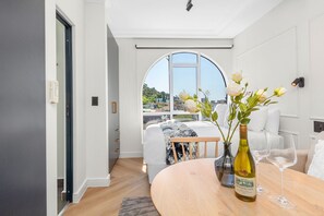 A modern hotel room with a large arched window, a bed with white linens, a wooden dining table with a wine bottle and glasses, and a vase with flowers. - The Valour 702 (Cape Town)