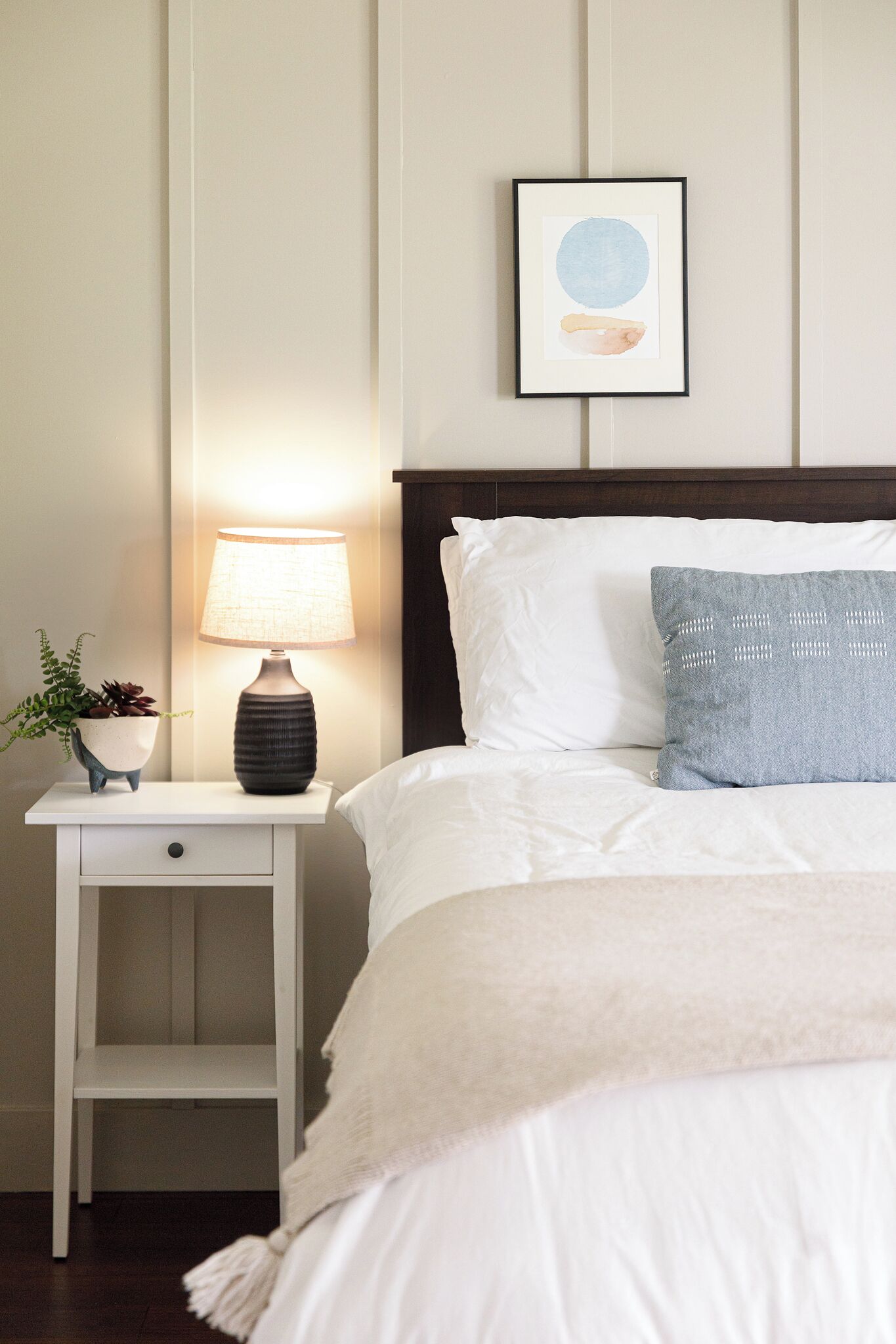 A neatly made bed with white bedding and a blue pillow, a bedside table with a lamp and a plant, and a framed picture on the wall.