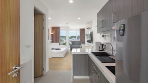 A modern kitchen with a built-in oven, a refrigerator, and a sink. There is a dining area with a table and chairs, and a bedroom with a bed and a window. - Studio in Ozone Condo in Kata A702 (Karon)