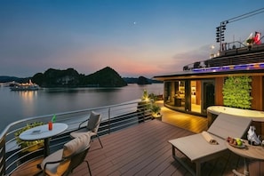 A deck with outdoor seating, a bar area, and a view of a river at dusk. - Calista Cruise - A Ha Long Bay Spectacle (Tuan Chau)