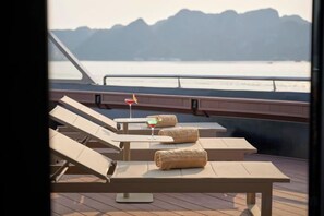 Eine Terrasse mit Liegestühlen und einem Tisch, mit Blick auf ein Gewässer und Berge.