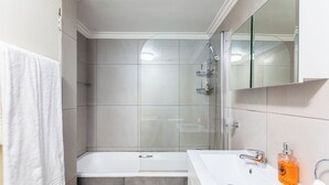 Et moderne badeværelse med en brusekabine i glas, et hvidt badekar og en vask med sæbedispenser.