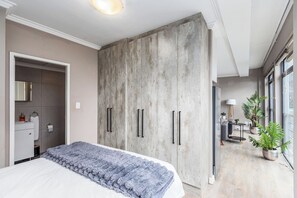 Una camera da letto con un ampio guardaroba, un letto con una coperta soffice, un bagno con uno specchio e una zona soggiorno con divano e piante.
