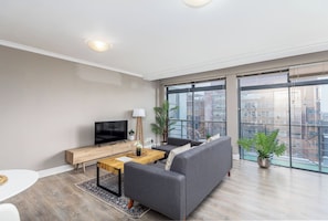 A modern living room with a flat-screen TV, a wooden coffee table, a grey sofa, and large windows offering a city view.