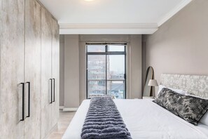 A bedroom with a large bed, a view of the city through a window, and a wardrobe with black handles.