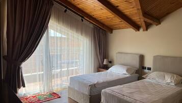 Una habitación con dos camas, vigas de madera en el techo, un ventanal con cortinas translúcidas y una alfombra roja con estampado.