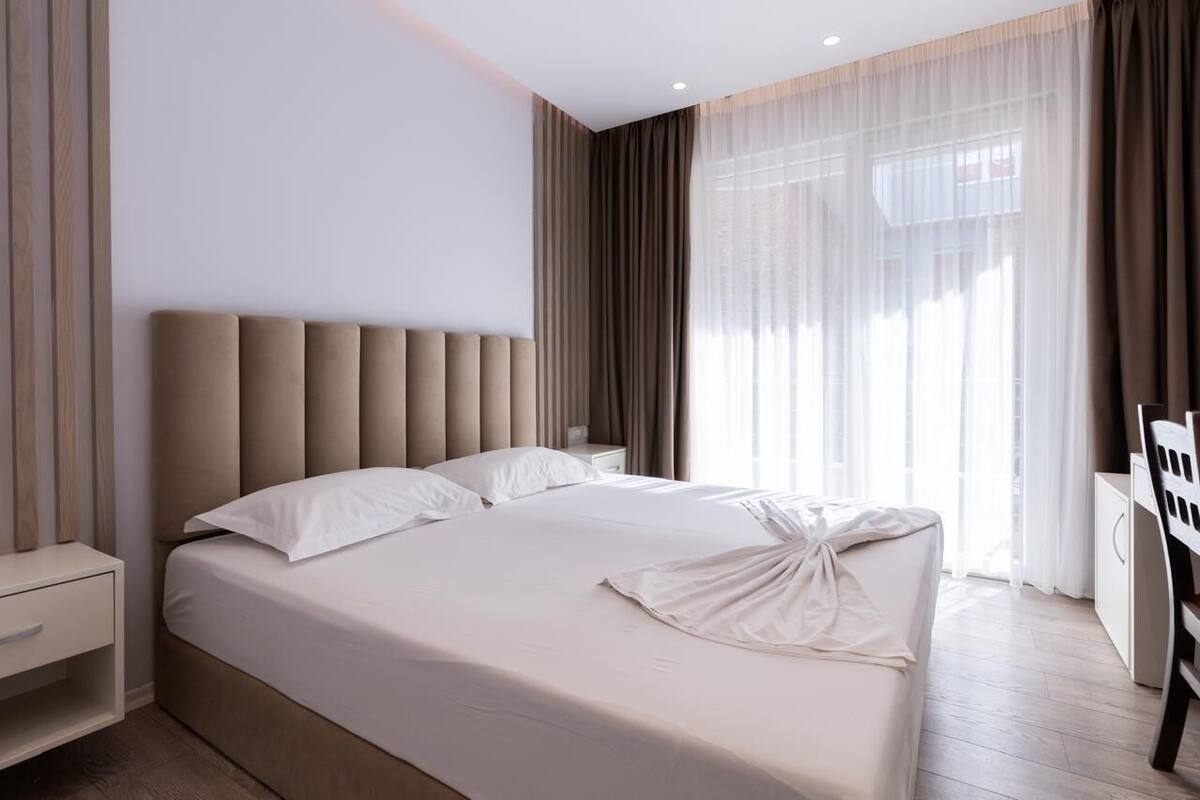 Une chambre d’hôtel moderne dotée d’un grand lit, d’une table de chevet, d’une armoire et offrant une vue sur l’extérieur à travers des rideaux transparents.