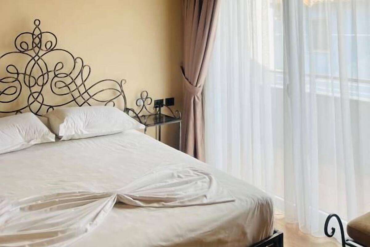 Une chambre avec un plafond en bois, un lit recouvert de draps blancs, une tête de lit en métal et une fenêtre ornée de rideaux légers.