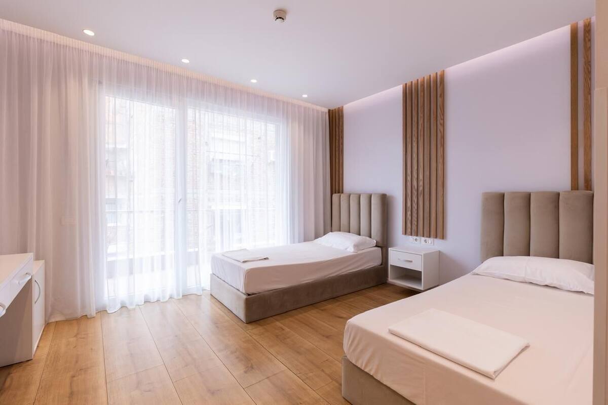 Une chambre d’hôtel moderne avec deux lits, un plancher en bois, une grande fenêtre avec des rideaux légers et un luminaire fixé au mur.