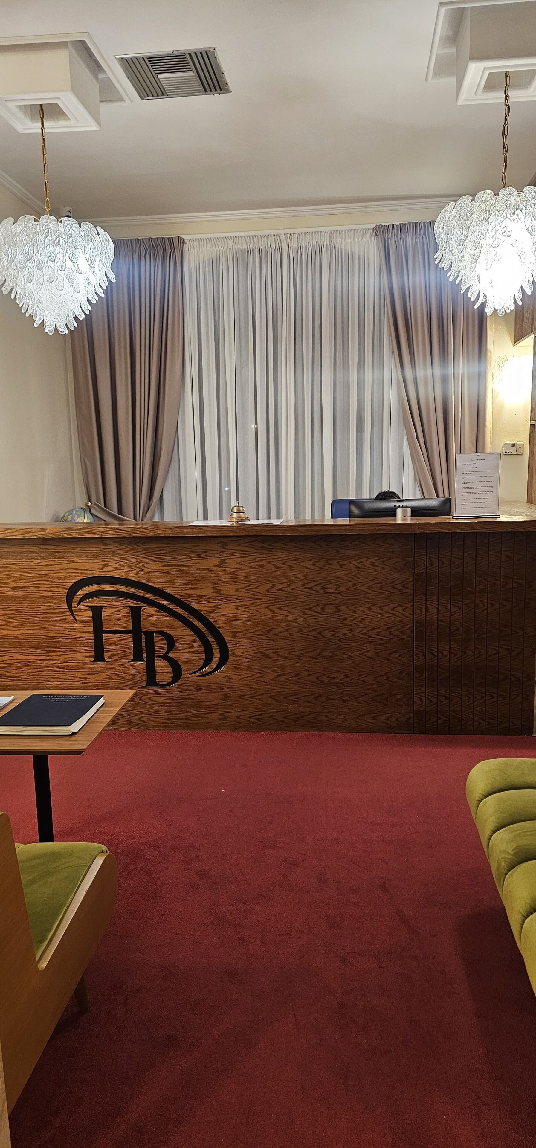 A hotel reception area with a wooden reception desk featuring a logo, a red carpet, and a green armchair.