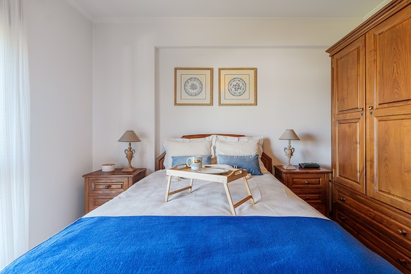 A bedroom with a bed, bedside tables, lamps, a wooden wardrobe, and two framed pictures on the wall.