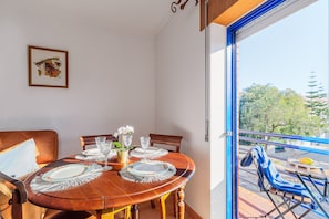 A dining area with a wooden table set for two, a framed picture on the wall, and a balcony with a chair.