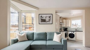 Ein modernes Wohnzimmer mit einem hellblauen Modulsessel, einem gerahmten Schwarz-Weiß-Foto an der Wand und Blick auf die Stadt durch große Fenster.