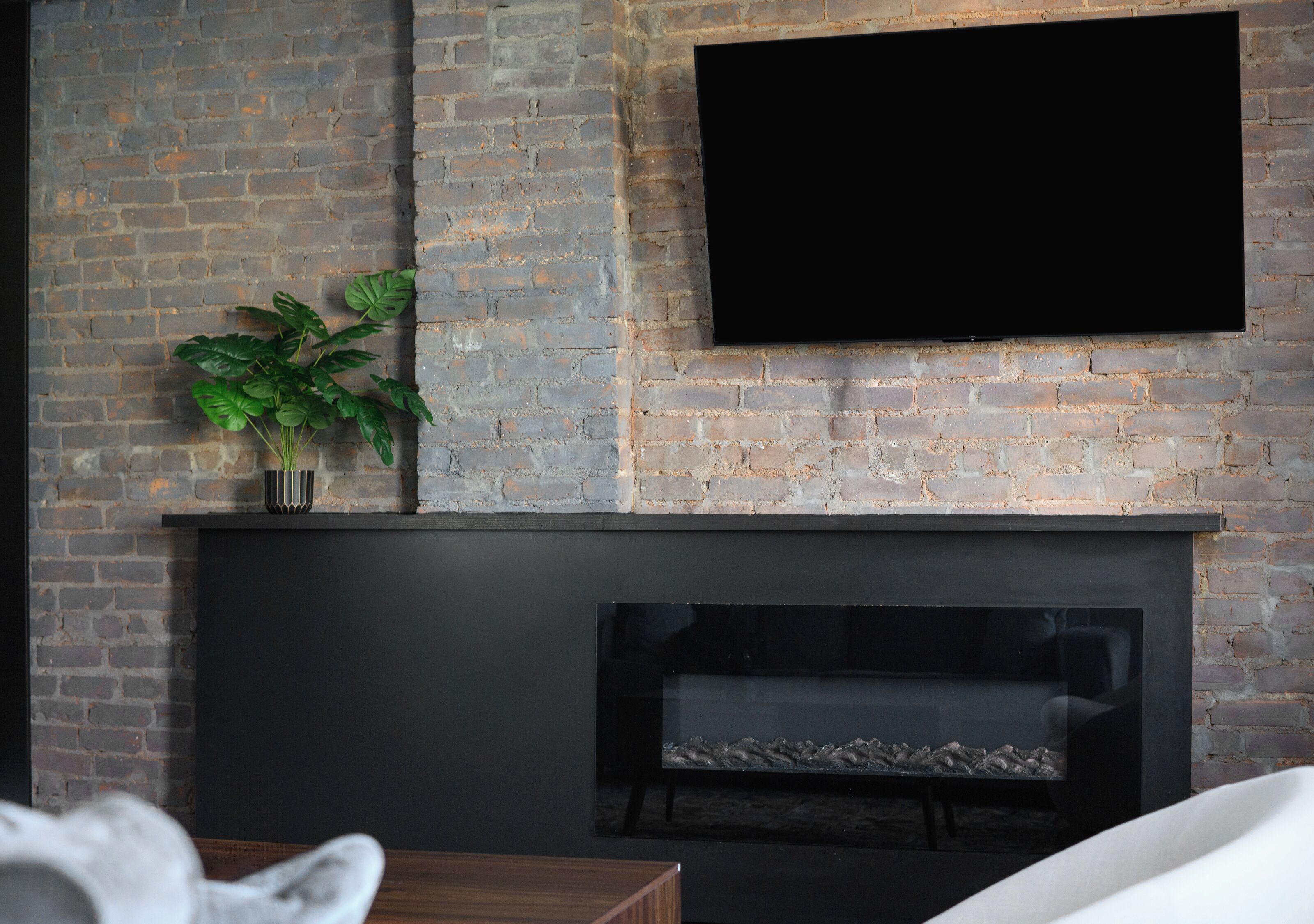A living room with a brick wall, a flat-screen TV mounted above a fireplace, and a potted plant on the mantel.