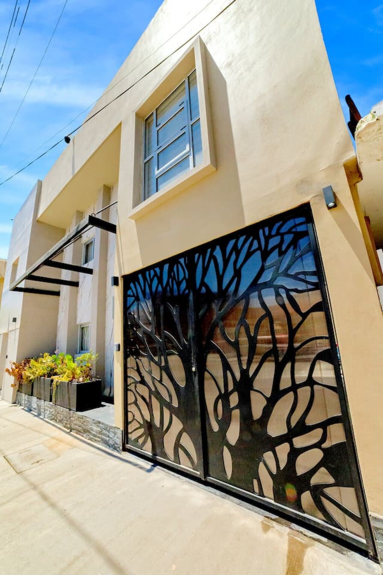 Une maison moderne à l’extérieur beige, avec un grand portail noir orné d’un motif d’arbre et une petite fenêtre au-dessus de la porte.
