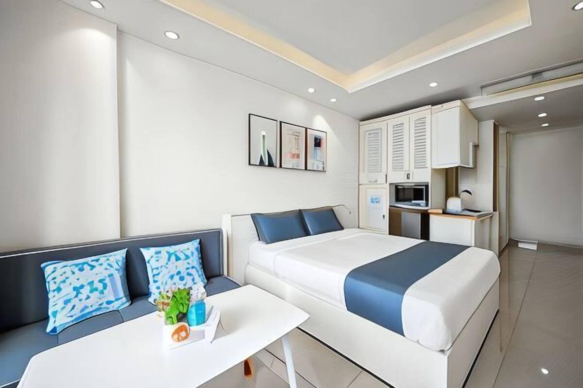 Ein modernes Hotelzimmer mit einem Bett, einem Sofa, einem kleinen Tisch mit Pflanze, einem Schreibtisch und einer Küchenzeile.