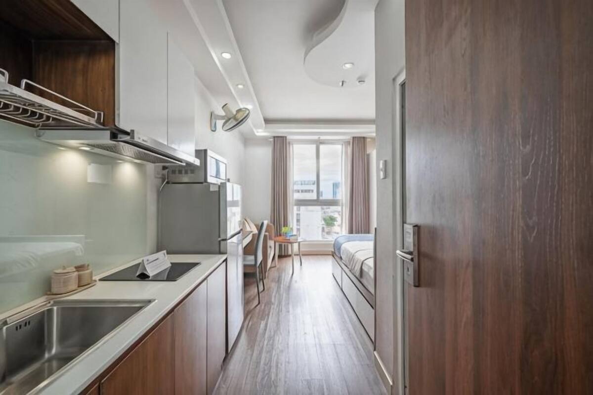 Ein kleines Hotelzimmer mit Küchenzeile, einem Bett, einem Schreibtisch und einem Fenster mit Vorhängen.