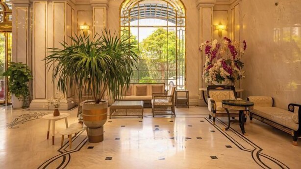 A spacious lobby with a large potted plant, a sofa, and a seating area with a view through a large arched window. - Cozrum Homes - Sonata Residence (Ho Chi Minh City)