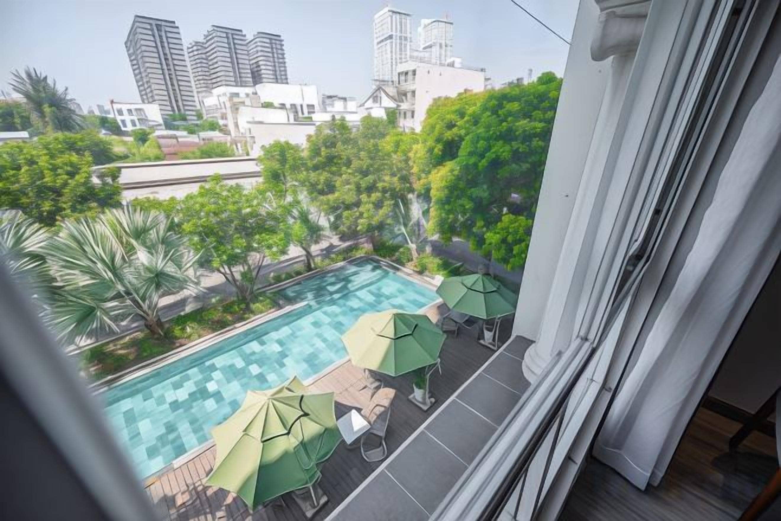 A rooftop pool with umbrellas and lounge chairs, surrounded by greenery and city buildings in the background.