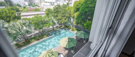 Una piscina en la azotea con sombrillas y sillas de descanso, rodeada de follaje y edificios de la ciudad al fondo.