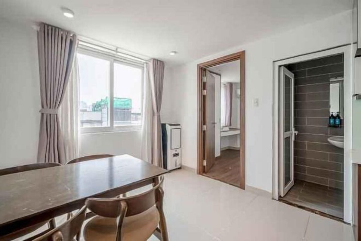 A room with a dining table, chairs, a window with curtains, a door leading to a bathroom, and a tiled floor.