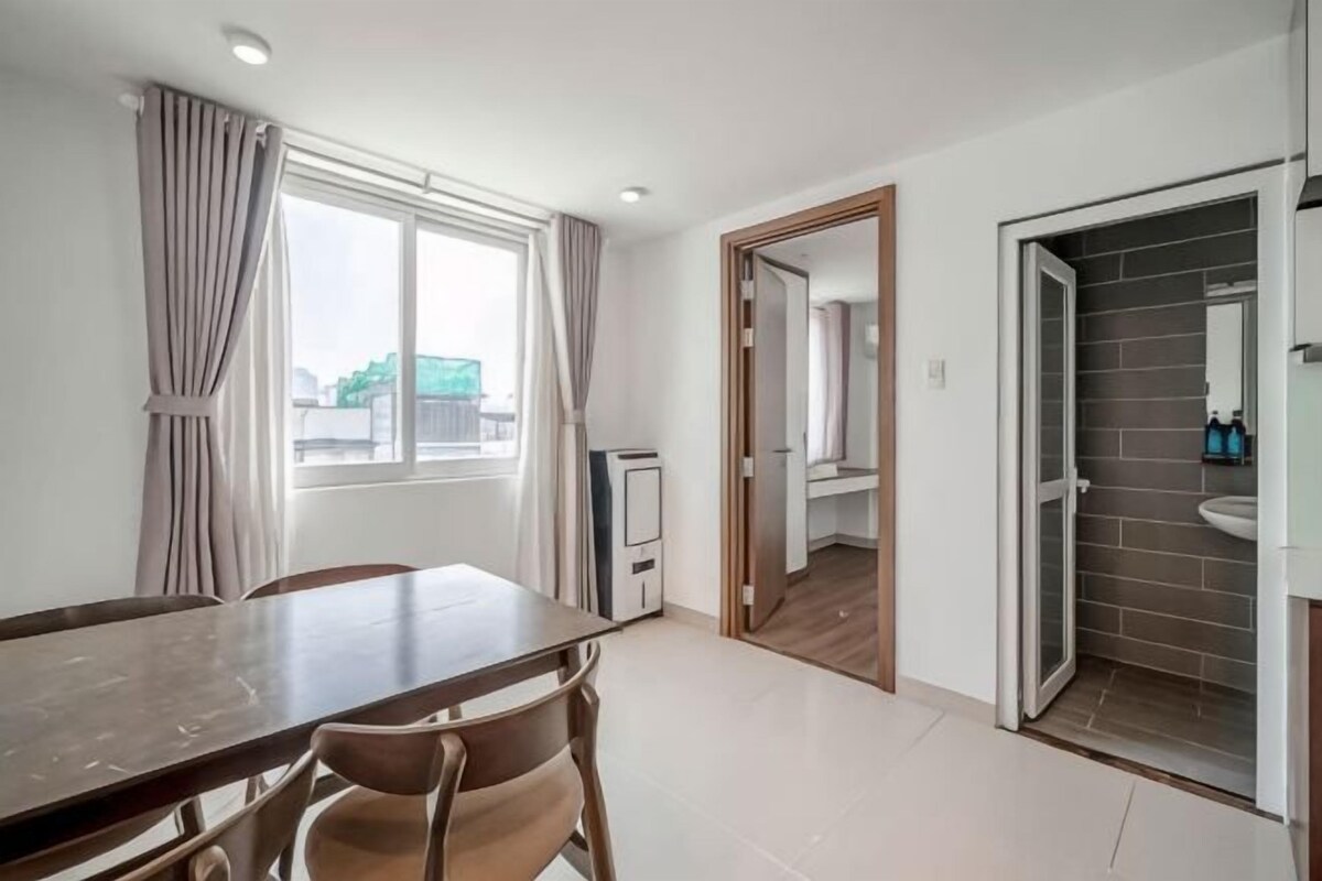 A room with a dining table, chairs, a window with curtains, a door leading to a bathroom, and a view of the outside.