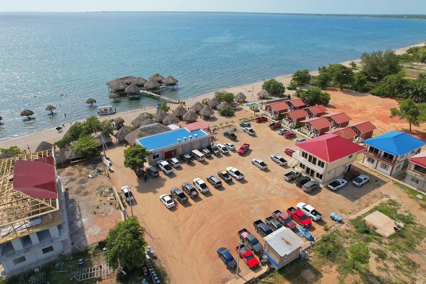 Vista aérea de una zona costera con playa, construcciones de techo de paja, un estacionamiento y varios edificios con tejados rojos.