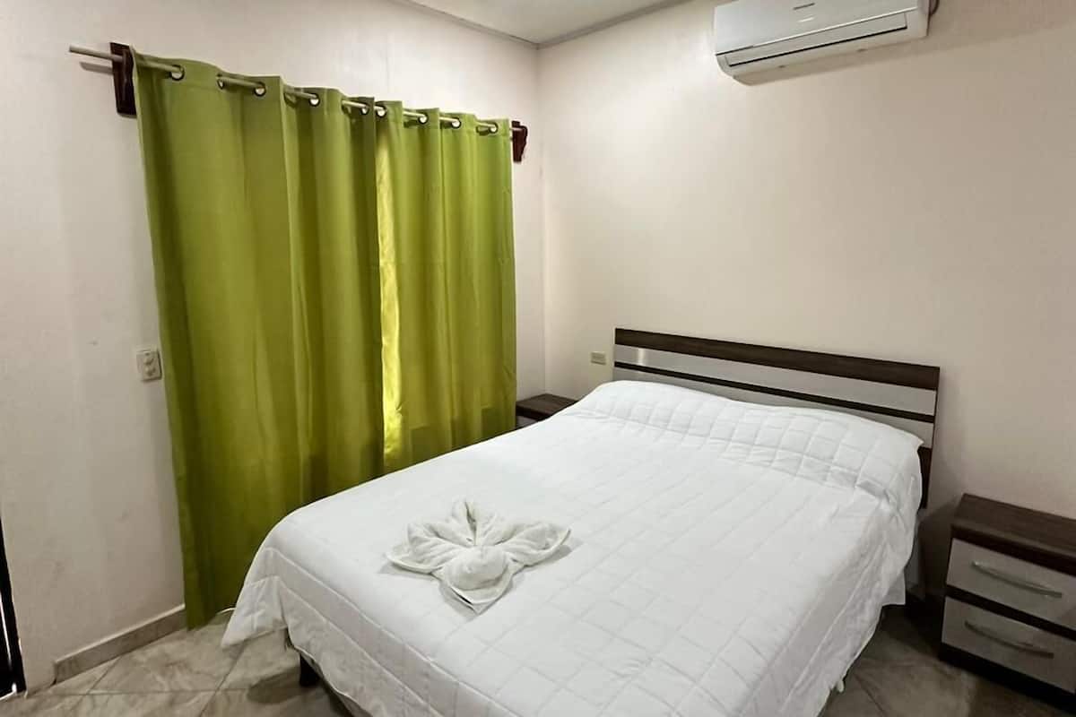 Una habitación de hotel con cama, mesita de noche, equipo de aire acondicionado y cortinas verdes.