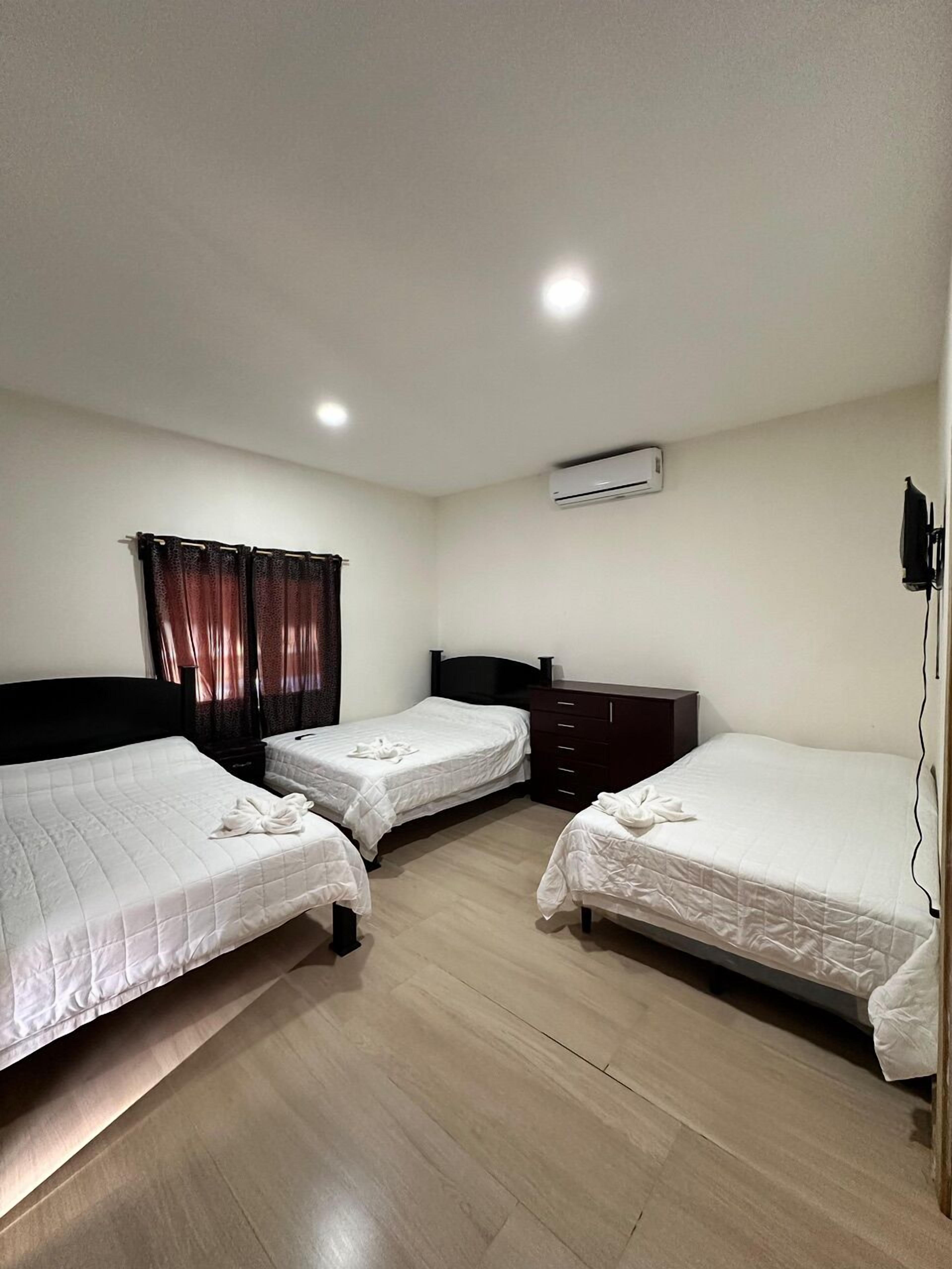 Habitación de hotel con tres camas, piso de madera, unidad de aire acondicionado y un armario de color oscuro.