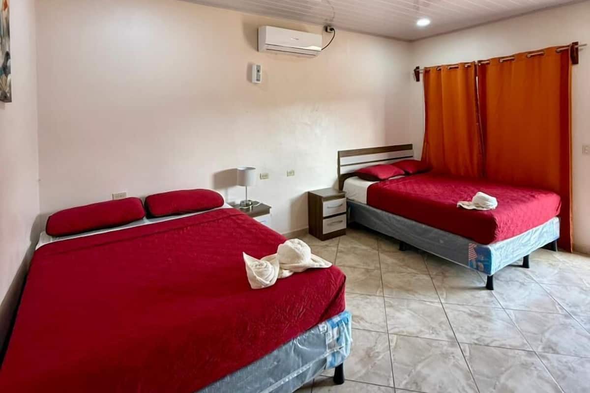 Habitación de hotel con dos camas, ventilador de techo, aire acondicionado, mesita de noche y ventana con cortinas.